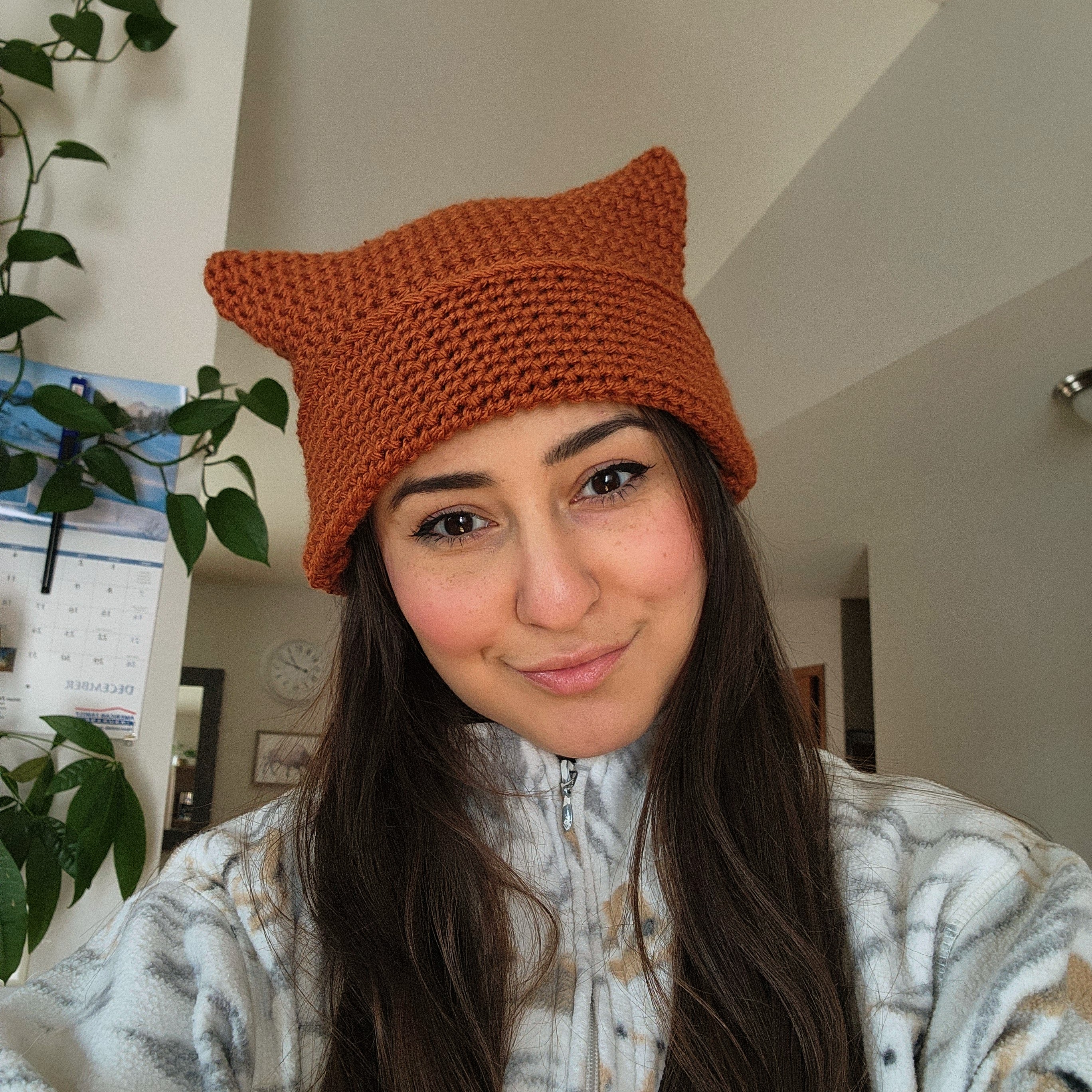 Keely Eagleshield, maker of the hat, long brown hair in a white jumper, wearing the burnt orange chunky beanie that resembles cat ears or a exaggerated corner look like a square - crocheted by hand